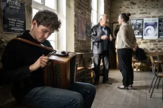 A young musician tuning an accordion in a rustic Breton hall, while an older local shows a dance step to a visitor in the background, bathed in soft late afternoon light.