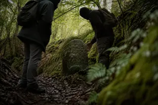 Two people observe a moss-covered stone marker along a shaded forest path, surrounded by damp earth and early greenery, in soft natural light.