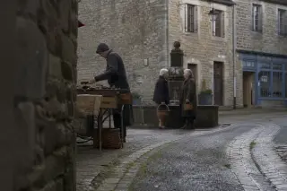 Early morning market setup in Bégard's historical center, side view of artisan by stone wall, two elderly locals chatting near granite fountain, soft natural light and candid, immersive mood.