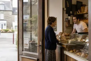Older woman speaking quietly to a young baker inside a rustic Breton bakery, with pastries and bread in the background and soft natural light highlighting the authentic, lived-in atmosphere.