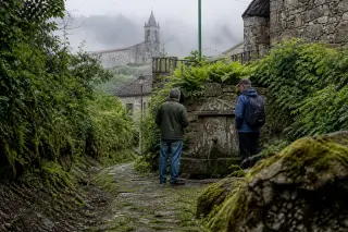 Two people quietly observing an old mossy fountain near a hamlet, with a chapel faintly visible in morning mist, captured at ground level after rain.