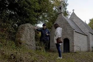 Deux personnes observent une chapelle bretonne en granit à l’ombre d’un bois, l’un montre une pierre gravée à l’autre, lumière douce en fin d’après-midi, ambiance discrète et réelle.