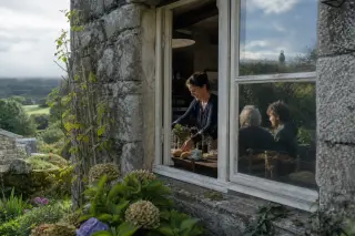 Local woman sets breakfast for two guests inside old Breton farmhouse, viewed through open window with mossy stone and soft morning light